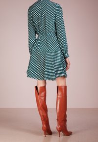 Grön klänning med vitt geometriskt mönster, långa ärmar och en utsvängd kjol. Matchad med knähöga, spetsiga, rostfärgade läderstövlar.