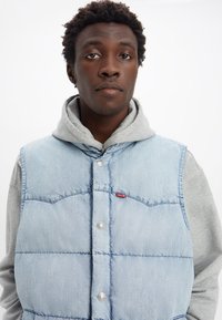Veste en denim bleu clair avec une texture matelassée, pressions en métal et un écusson rouge, superposée à un hoodie gris.