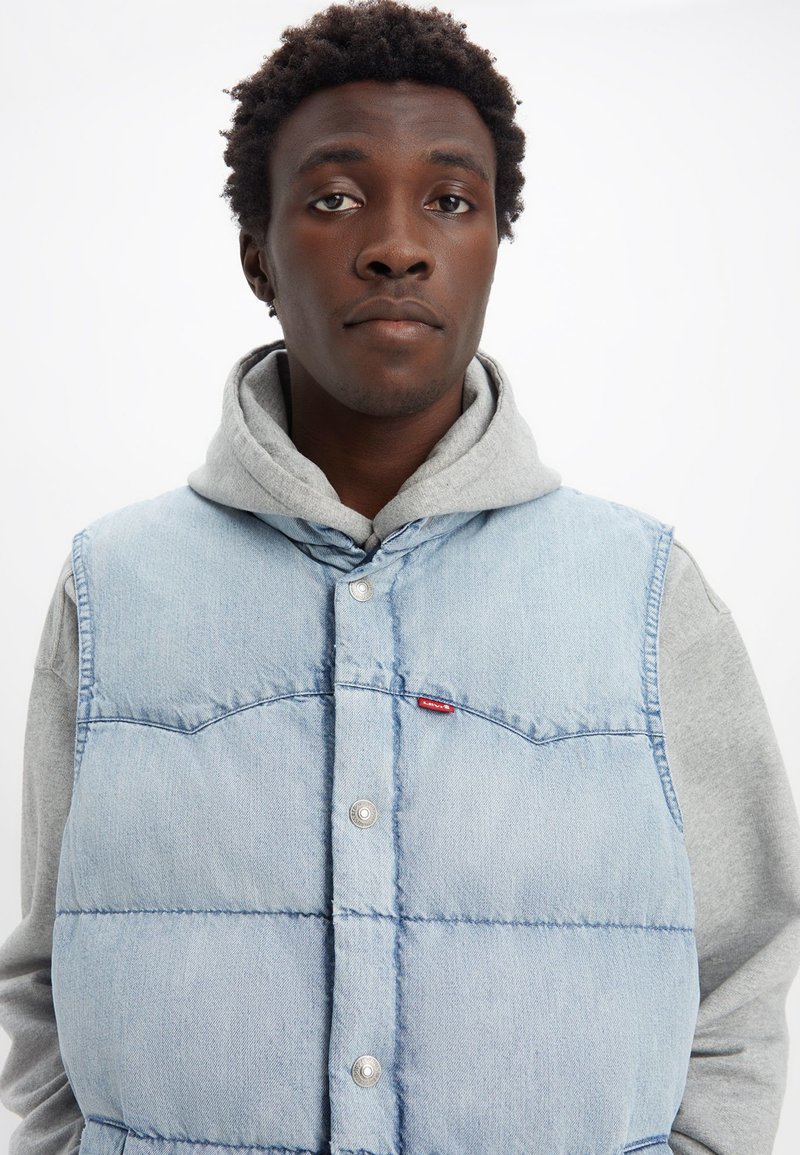 Veste en denim bleu clair avec une texture matelassée, pressions en métal et un écusson rouge, superposée à un hoodie gris.