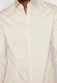 Cream-colored button-up shirt made from smooth cotton fabric. Features a classic collar and six buttons down the front with a minimal design.