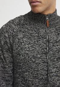 Textured zip-up sweater in black and gray with a high collar and subtle pattern, featuring a leather tag on the zipper pull.