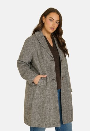 Donna che indossa un lungo cappotto grigio a spina di pesce sopra un maglione scuro e jeans blu, con una mano nella tasca del cappotto, su uno sfondo semplice.