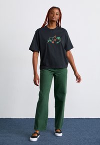 Black cotton t-shirt with graphic design in green and red, paired with green straight-leg pants and black slip-on shoes with white soles.