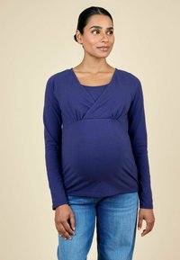 Top de maternidad de manga larga en color púrpura oscuro con escote en V y detalles fruncidos debajo del busto, combinado con jeans de color azul claro. Textura de tela suave.