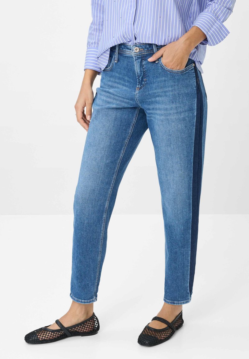 Donna che indossa jeans blu con una striscia scura laterale, una maglietta a righe blu e scarpe Mary Jane in rete nere, in piedi con una mano in tasca.