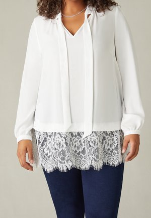 Kvinde iført en hvid langærmet bluse med V-udskæring, sløjfe og blondekant kombineret med mørkeblå bukser.