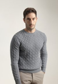 Suéter gris de punto trenzado con mangas largas y dobladillo acanalado. La tela presenta un patrón texturizado y elevado. Llevado con pantalones beige claros.