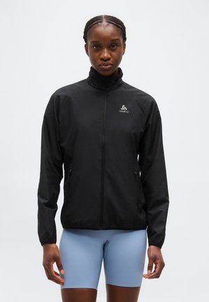 Veste noire à fermeture éclair avec col haut, logo sur la poitrine, portée par une personne aux cheveux tressés ; associée à un short de cyclisme bleu clair.