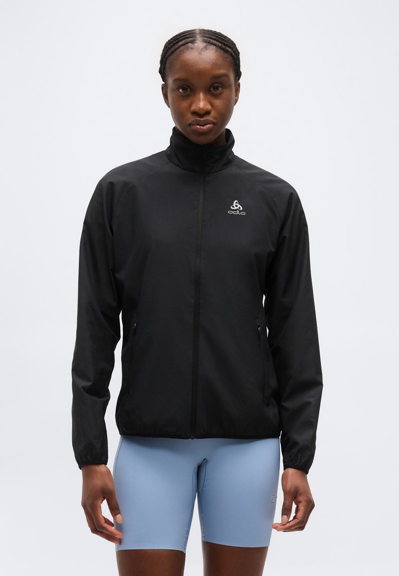 Veste noire à fermeture éclair avec col haut, logo sur la poitrine, portée par une personne aux cheveux tressés ; associée à un short de cyclisme bleu clair.