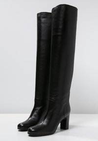 Bottes noires en cuir montantes avec une finition brillante, bout rond et talon bloc. Texture lisse avec des détails de couture minimaux.