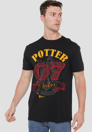 Mann trägt ein schwarzes T-Shirt mit gelbem Text "Potter,", roter Nummer "07," und "Seeker" über einem Gryffindor-Wappen Design, schaut seitlich.