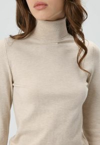 Pull à col roulé beige en tissu côtelé doux. Coupe ajustée avec un col haut et une texture lisse, idéal pour superposer.