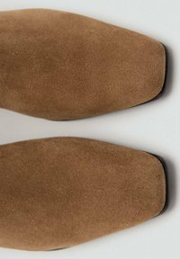 Botas de tobillo de ante marrón con punta afilada, textura suave y una silueta elegante. Los detalles de la suela negra añaden contraste.