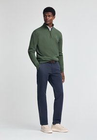 Grön herrtröja i stickad kvalitet med dragkedja i kragen, ihop med marinblå chinos och ljusbeiga sneakers för en avslappnad look.