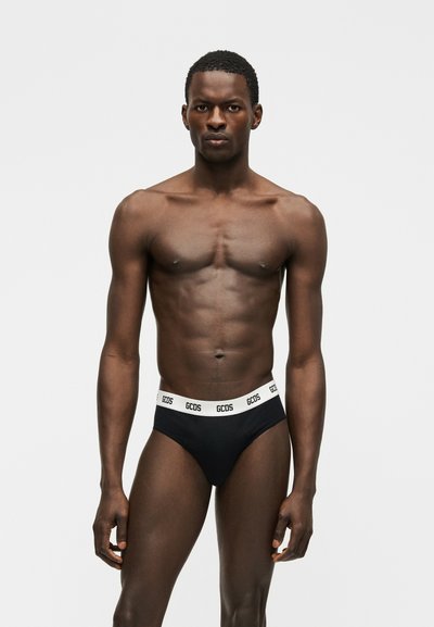 Άνδρας μοντέλο στέκεται μπροστά σε απλό φόντο φορώντας μαύρα εσώρουχα με λευκή ελαστική ζώνη μέσης με το λογότυπο GCDS, κοιτάζοντας μπροστά με ουδέτερη έκφραση.