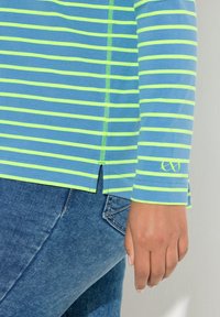 Käsi lepää sivulla, pukien sinikeltaraidallisen pitkähihainen paidan, jossa on pieni sivuhalkio, yhdistettynä sinisiin farkkuihin.