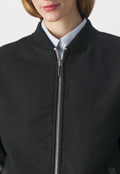 Melns bomber stils apvalks ar rāvējslēdzēju priekšpusē, apaļu apkakli un teksturētu audumu. Izteiksmīgs sudraba rāvējslēdzējs piešķir praktisku detaļu.