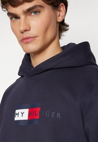 Mornarsko plava jakna s kapuljačom s prednjim logotipom koji sadrži pravokutne akcentne boje u crvenoj, bijeloj i plavoj. Natpis glasi "TOMMY HILFIGER" u svijetlosivoj boji. Mekani teksturirani materijal.