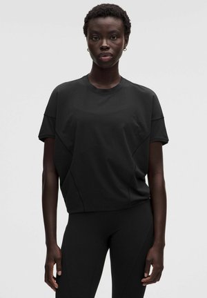 Donna con capelli corti e ricci che indossa una maglietta nera oversize e pantaloni neri aderenti, in piedi davanti a uno sfondo grigio semplice.