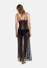 Longue robe noire transparente en dentelle avec bretelles fines réglables, ornée de motifs floraux et de fentes latérales, portée par-dessus des sous-vêtements noirs.