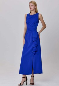 Femme portant une robe maxi sans manches bleu royal avec des volants, une fente sur le côté, et des sandales à talons noirs, se tenant contre un fond uni.