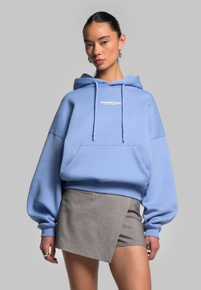 Femme portant un sweat à capuche oversize bleu clair avec un texte blanc et une mini-jupe asymétrique grise sur fond gris uni.