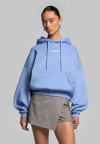 Lys blå oversized hoodie lavet af blødt stof, med en frontlomme og snørrefunktion i hætten, parret med en grå asymmetrisk miniskørt.