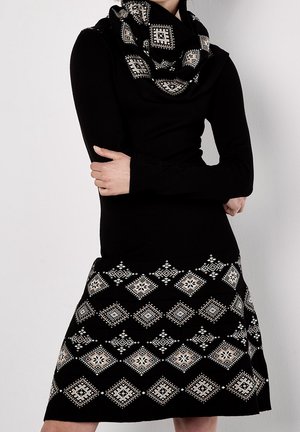 Femme portant une robe noire à manches longues et une écharpe avec des motifs géométriques blancs en forme de losange, les bras croisés, debout devant un fond clair uni.