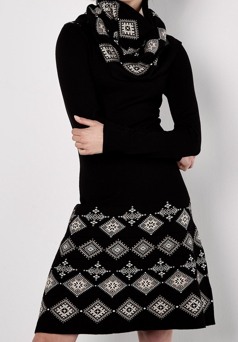 Femme portant une robe noire à manches longues et une écharpe avec des motifs géométriques blancs en forme de losange, les bras croisés, debout devant un fond clair uni.