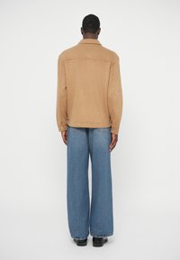 Tan wool-blend shirt jacket with a collar, back yoke, and buttoned cuffs, paired with loose-fitting blue denim jeans and black shoes.