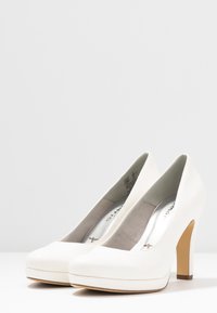 Pár bílých vysokých platformových lodiček s béžovými podpatky na bílé ploše proti jednoduchému světle šedému pozadí.