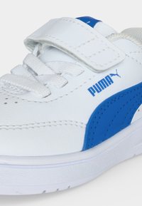 Λευκό αθλητικό παπούτσι Puma με μπλε ρίγα, λουράκι με βέλκρο και κορδόνια, που δείχνει πλευρικές και μπροστινές λεπτομέρειες σε απλό φόντο.