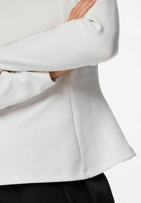 Bijela majica dugih rukava od mekog, glatkog materijala opuštenog kroja, s laganim širem rubom i zaobljenim manžetama.