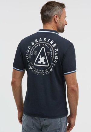 Mann trägt ein marineblaues Poloshirt mit weißem und blauem Kragenbesatz und nautischem Logo auf der Rückseite, kombiniert mit grauen Shorts.