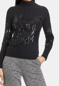 Pull noir côtelé à col montant, orné de sequins sur le devant et les manches. Associé à un pantalon gris à motifs.