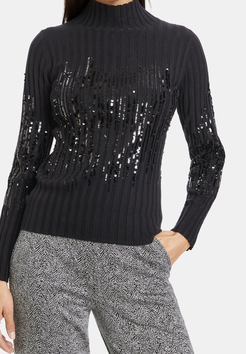 Pull noir côtelé à col montant, orné de sequins sur le devant et les manches. Associé à un pantalon gris à motifs.