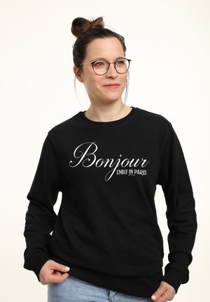 Frau mit Brille und Dutt-Frisur trägt einen schwarzen Pullover mit der Aufschrift „Bonjour Emily in Paris“ und hält den Saum vor einem schlichten Hintergrund.