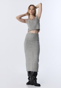 Kobieta ubrana w szary bezrękawnik z dzianiny i pasującą długą spódnicę, do której dobrała czarne kozaki za kolano, pozująca z uniesionymi ramionami.