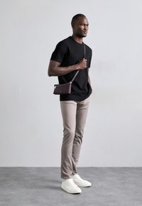 Sort tekstureret T-shirt, beige slim-fit bukser, hvide sneakers og en lille brun crossover-taske med lynlås. Står foran en neutral baggrund.