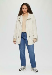 Lichtbeige jas met een hoge kraag, twee voorkruiden en een verstelbaar trekkoord. Draag het over een beige sweater en blauwe jeans. Zilveren sneakers.