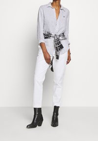 Person som bär svarta ankelskor, vita byxor, en vit randig skjorta med knappar, och en svart-vit rutmönstrad skjorta knuten runt midjan.