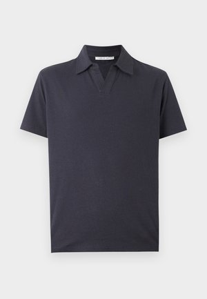 Homme debout, mains dans les poches, portant un polo noir à manches courtes, un short bleu clair, des chaussettes blanches et des baskets beiges sur fond blanc.