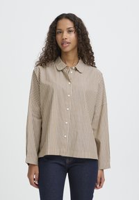 Kvinde med krøllet hår iført en beige og hvid vertikalstribet skjorte med knapper og mørkeblå jeans, stående foran en ensfarvet baggrund.