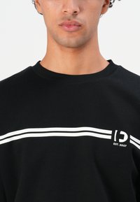 Young man wearing a black sweatshirt with two white stripes and a partial logo with "EST. 2007" printed on the chest.