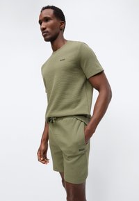 Άνδρας που φοράει ελαιί πράσινο κοντομάνικο πουκάμισο με υφή και αντίστοιχα σορτς με λογότυπα "BOSS", στέκεται με το ένα χέρι στην τσέπη.