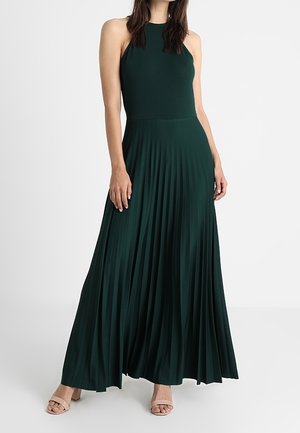 Woman wearing a sleeveless, dark green pleated maxi dress with a high neckline and beige open-toe heels, standing against a plain background.