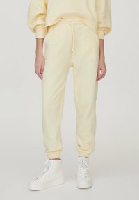Jasnożółte spodnie dresowe z miękkiego materiału, z elastycznym paskiem w talii ze sznurkiem, zwężanymi mankietami i bocznymi kieszeniami, w zestawie z białymi sneakersami.