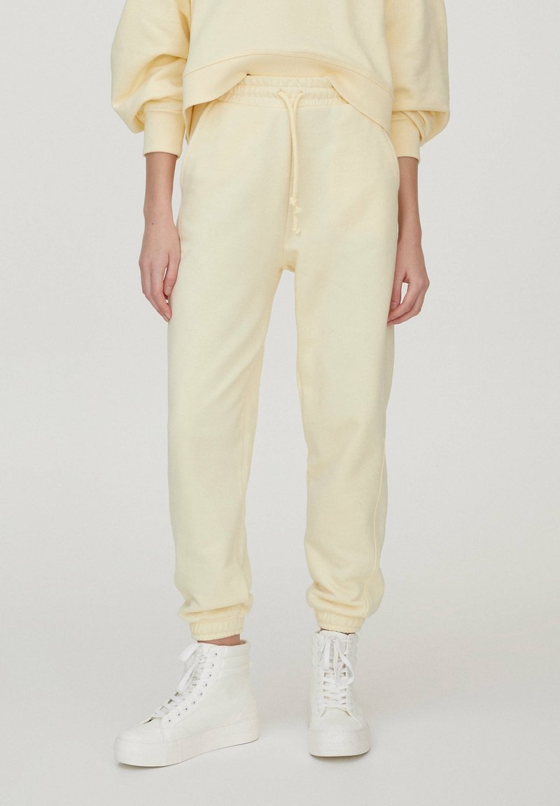 Jasnożółte spodnie dresowe z miękkiego materiału, z elastycznym paskiem w talii ze sznurkiem, zwężanymi mankietami i bocznymi kieszeniami, w zestawie z białymi sneakersami.