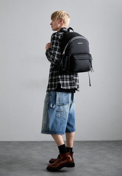 Rucsac negru din material texturat, cu detalii din fermoar argintii, purtat peste o cămașă în carouri și asortat cu pantaloni scurți din denim bleu deschis și cizme maronii.