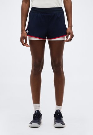 Športnik, ki nosi mornarsko modre večplastne tekaške kratke hlače z rdečim robom, bele nogavice do gležnja in črne športne copate na nevtralnem ozadju.